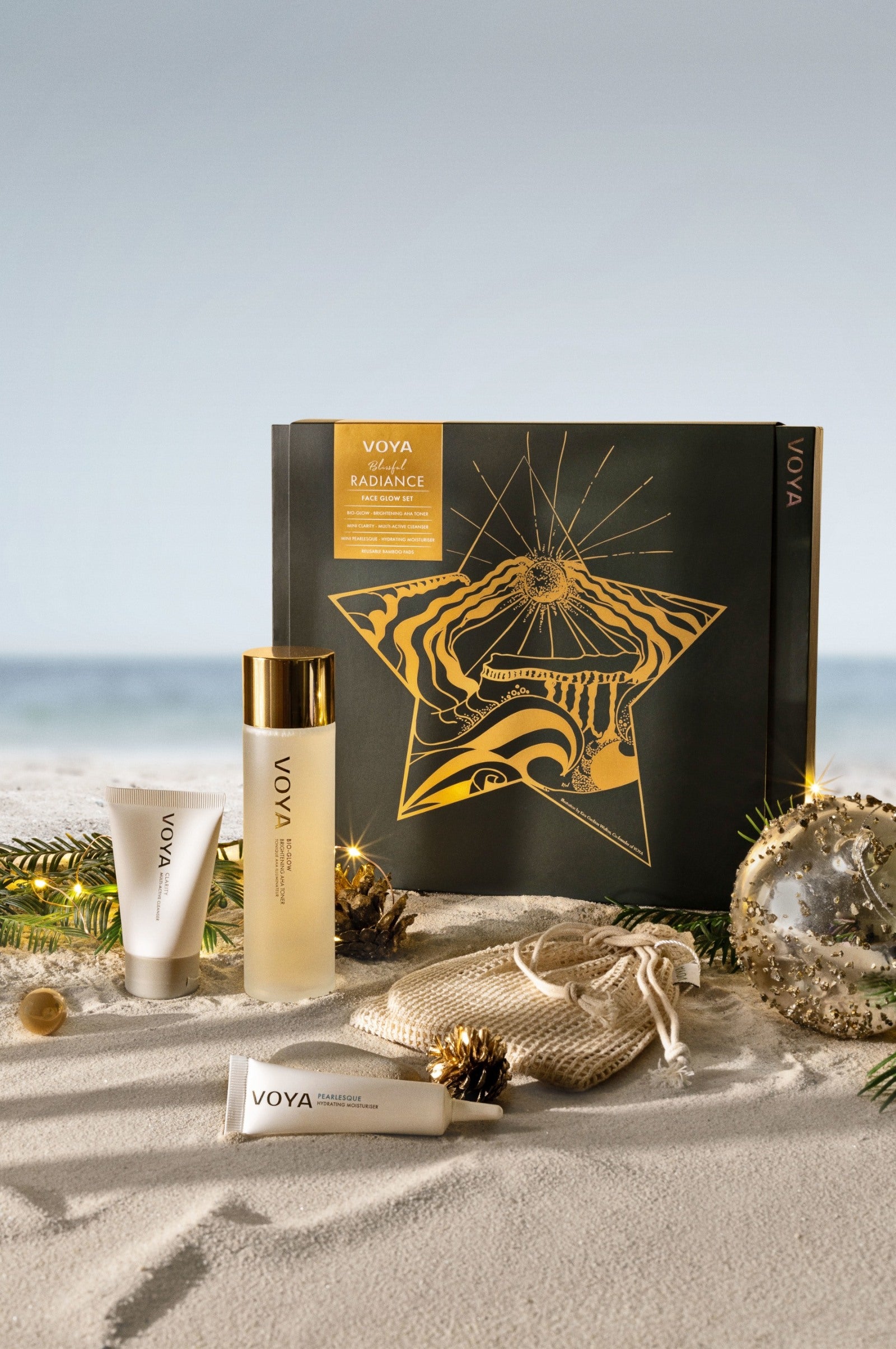 VOYA Blissful Radiance Facial Gift Set containing Bio-Glow AHA Toner, Mini Clarity Cleanser, Mini Pearlesque Moisturiser and three reusable bamboo pads against a beach backdrop. 